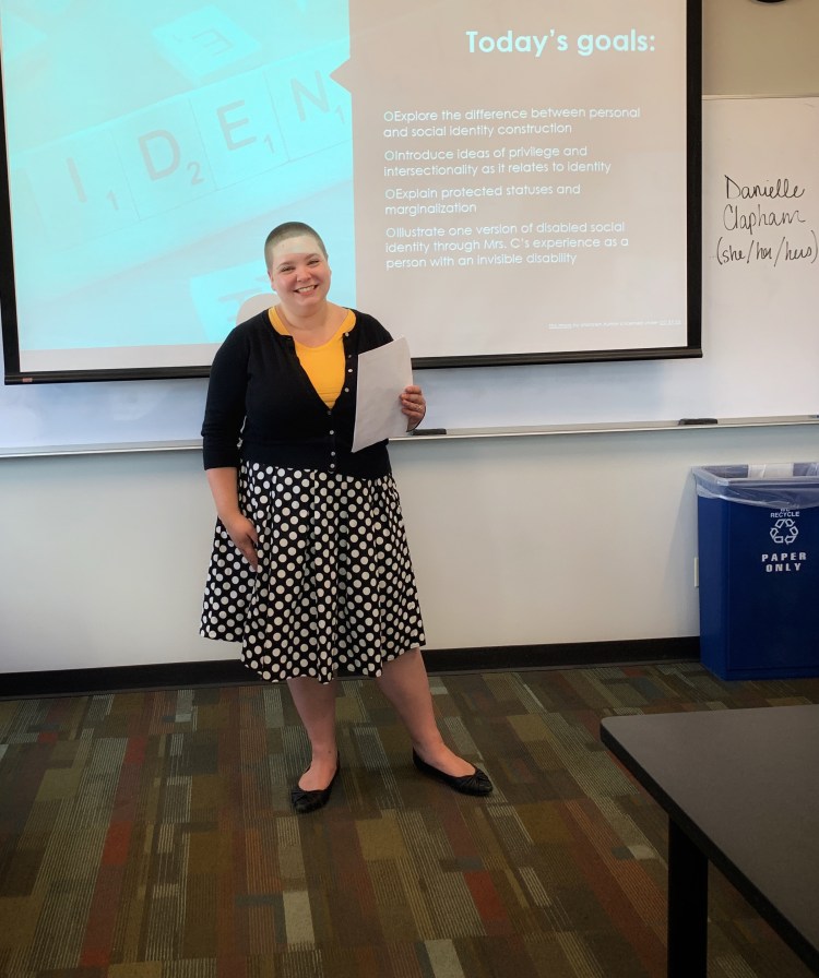 Danielle Clapham in polka dot skit, yellow shirt, and black cardigan holding a piece of paper and standing in front a powerpoint screen.